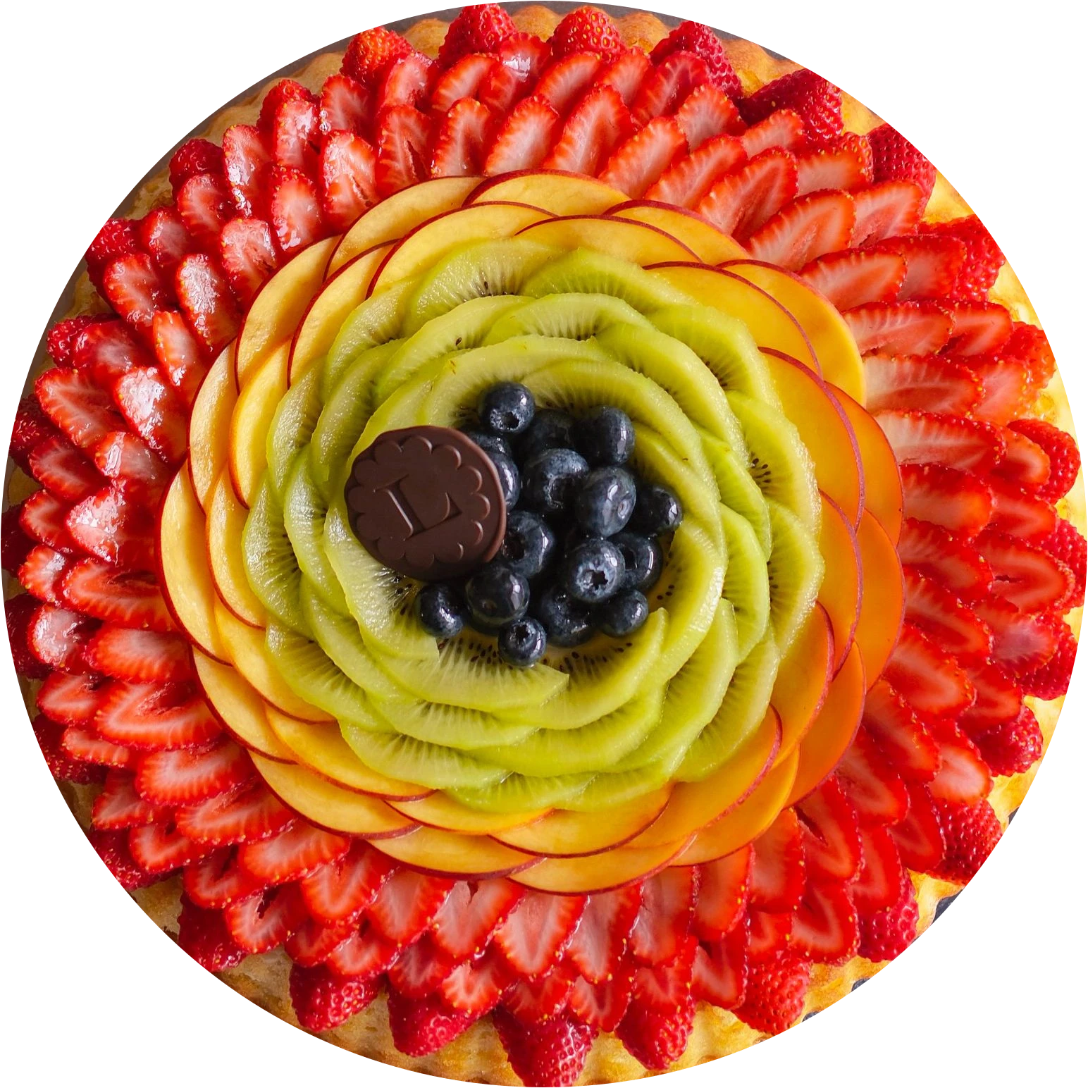 Tarta flor de Lampone vista desde arriba: fresas, mango y kiwi en pétalos alrededor de arándanos y una medalla de chocolate con la L