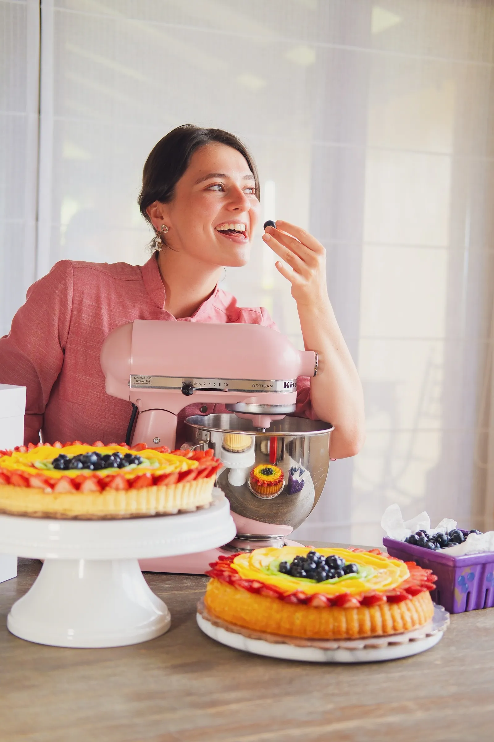 La pastelera de Lampone hoy, sonriendo junto a su batidora rosada y tres tartas de fruta